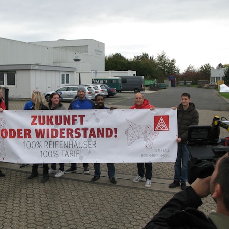 Acht Männer und zwei Frauen vor einem Werkstor mit einem Protest-Transparent der IG Metall. Rechts im Vordergrund steht ein Fernseh-Kameramann.