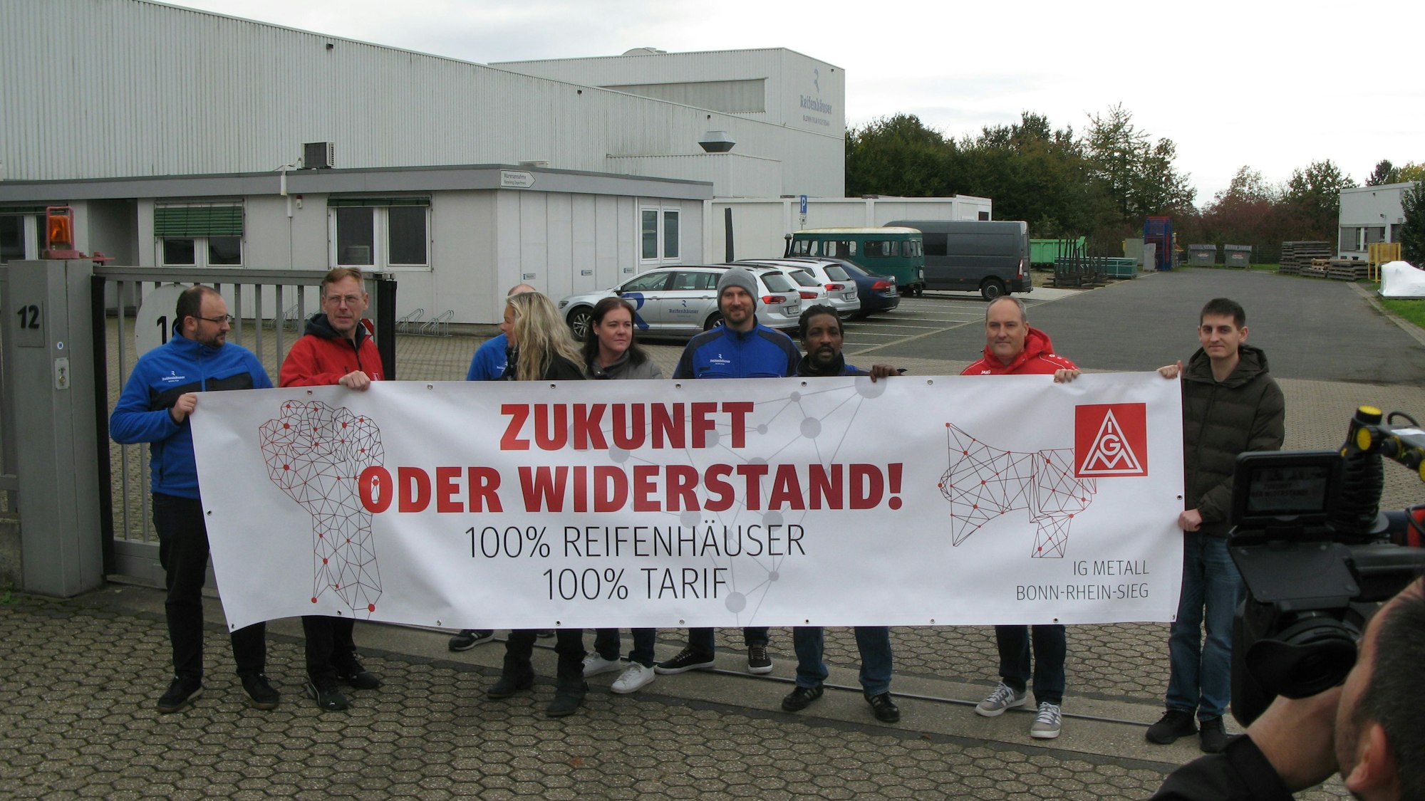 Acht Männer und zwei Frauen vor einem Werkstor mit einem Protest-Transparent der IG Metall. Rechts im Vordergrund steht ein Fernseh-Kameramann.