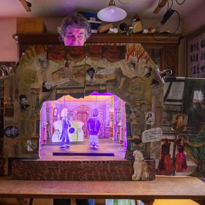 Ein Mann steht hinter einem Papiertheater, das eine Szene einer Komödie zeigt.