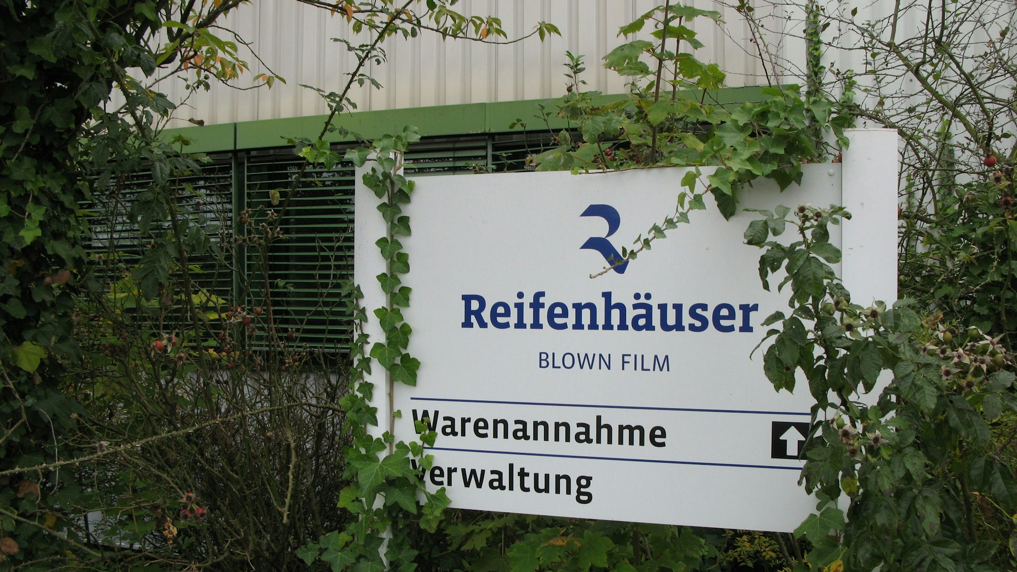 Ein Firmenschild des Unternehmens Reifenhäuser vor einer Halle, das zum Teil von Pflanzen überwuchert ist.
