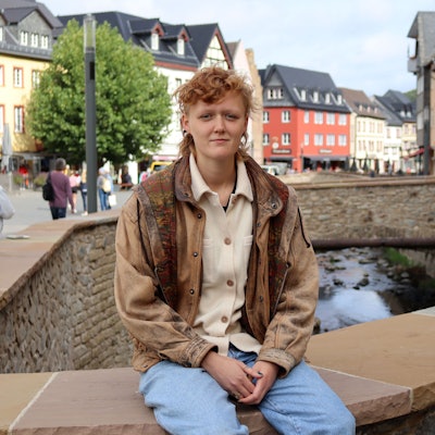 Svea Kirschmeier sitzt auf der Erftmauer in Bad Münstereifel. Sie trägt Jeans und eine braune Lederjacke.