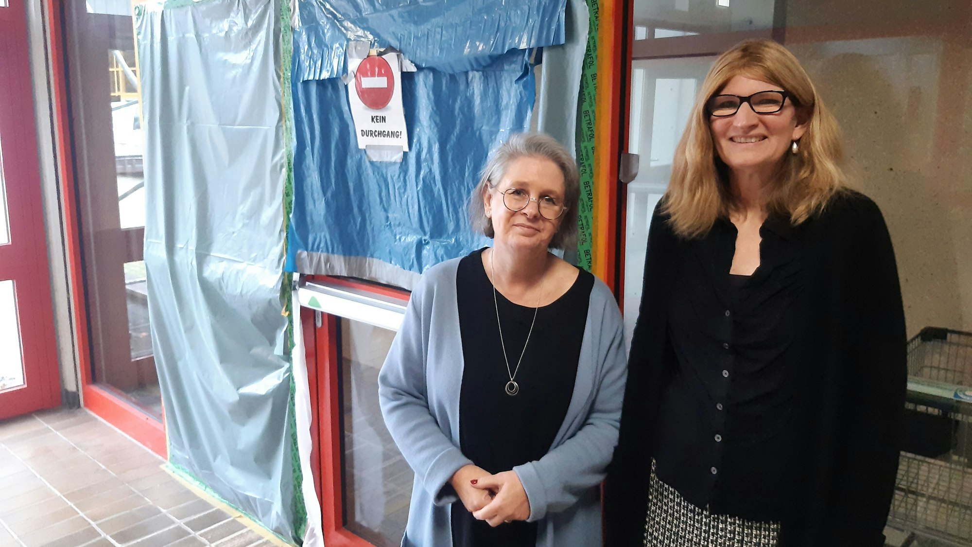 Das Bild zeigt zwei Frauen vor einem mit einer Plane abgesperrten Bereich in einer Schule.