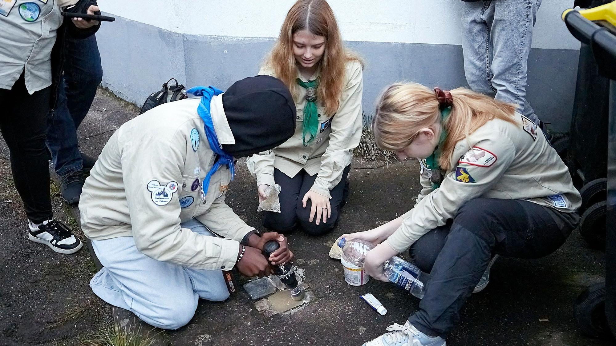 Drei Jugendliche in Pfadfinderhemden hocken auf dem Boden. Sie reinigen mehrere Stolpersteine, mit denen an von den Nationalsozialisten ermordete Mitbürger erinnert wird.
