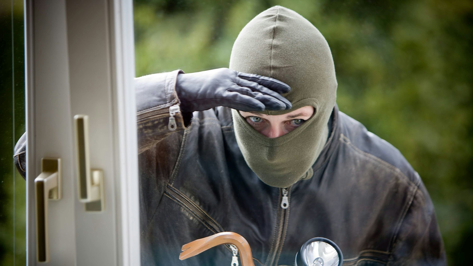Das Symbolbild zeigt einen maskierten Mann mit Einbruchswerkzeug hinter der Scheibe eines gekippten Fensters.