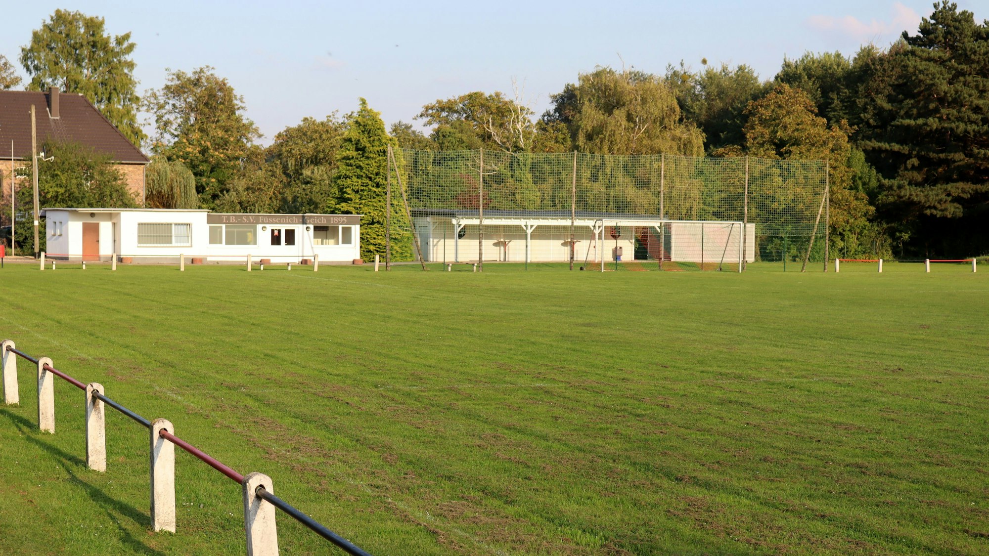 Das Bild zeigt den Rasenplatz mit Umkleidegebäuden und Bäumen im Hintergrund.