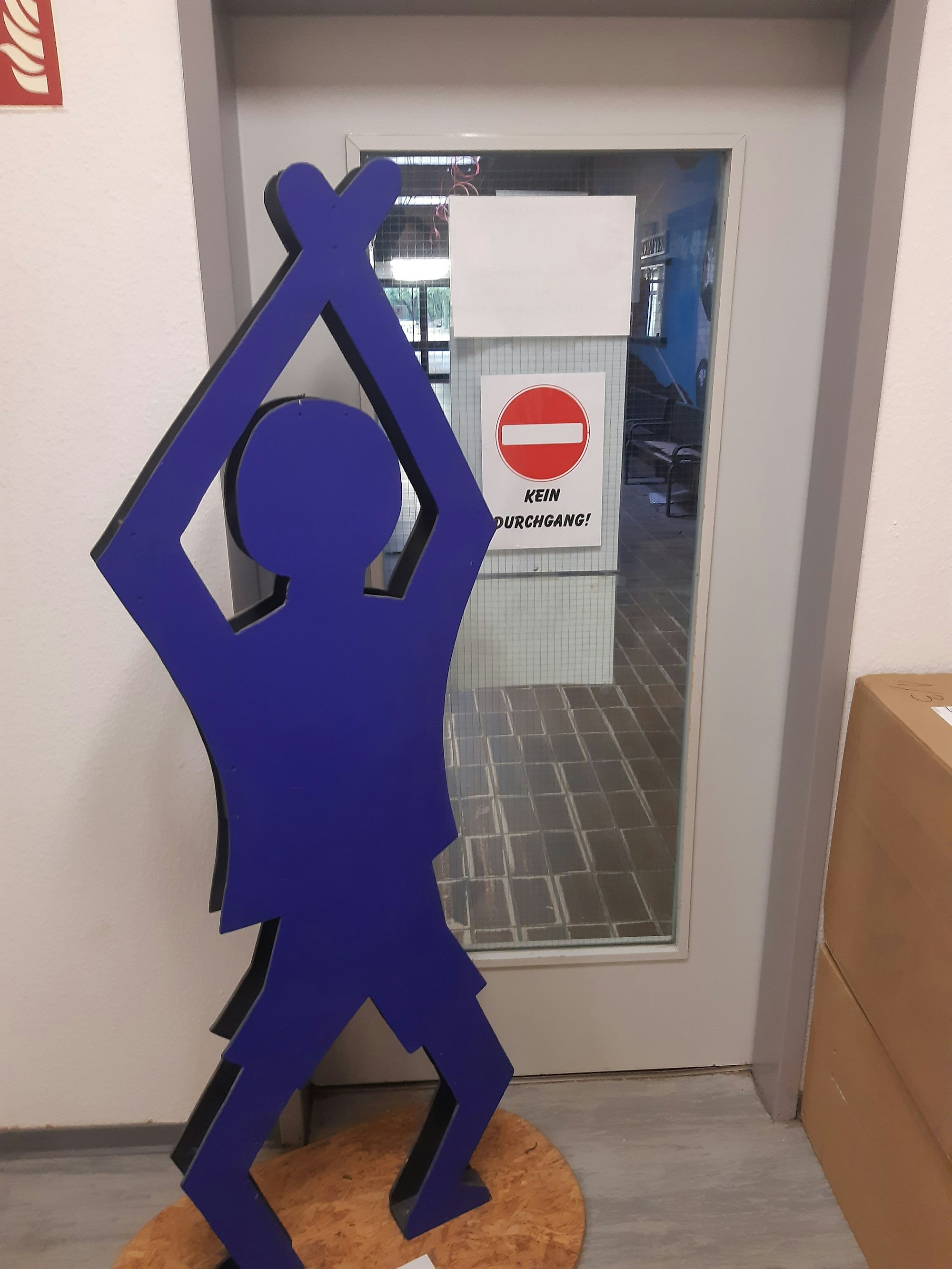 Das Bild zeigt einen blauen Pappaufsteller in Form eines Männchens vor einer Tür in einer Schule.