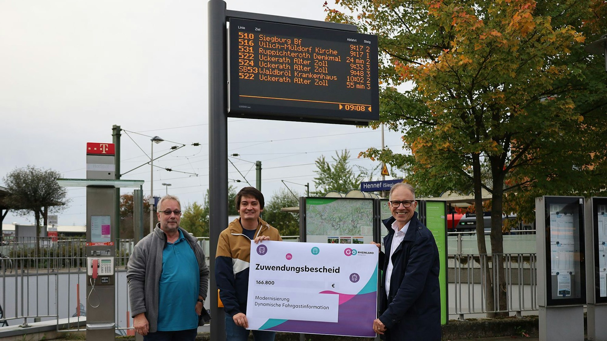 Drei Männer halten einen Zuwendungsbescheid, im Hintergrund sieht man die Anzeigetafel einer Haltestelle.