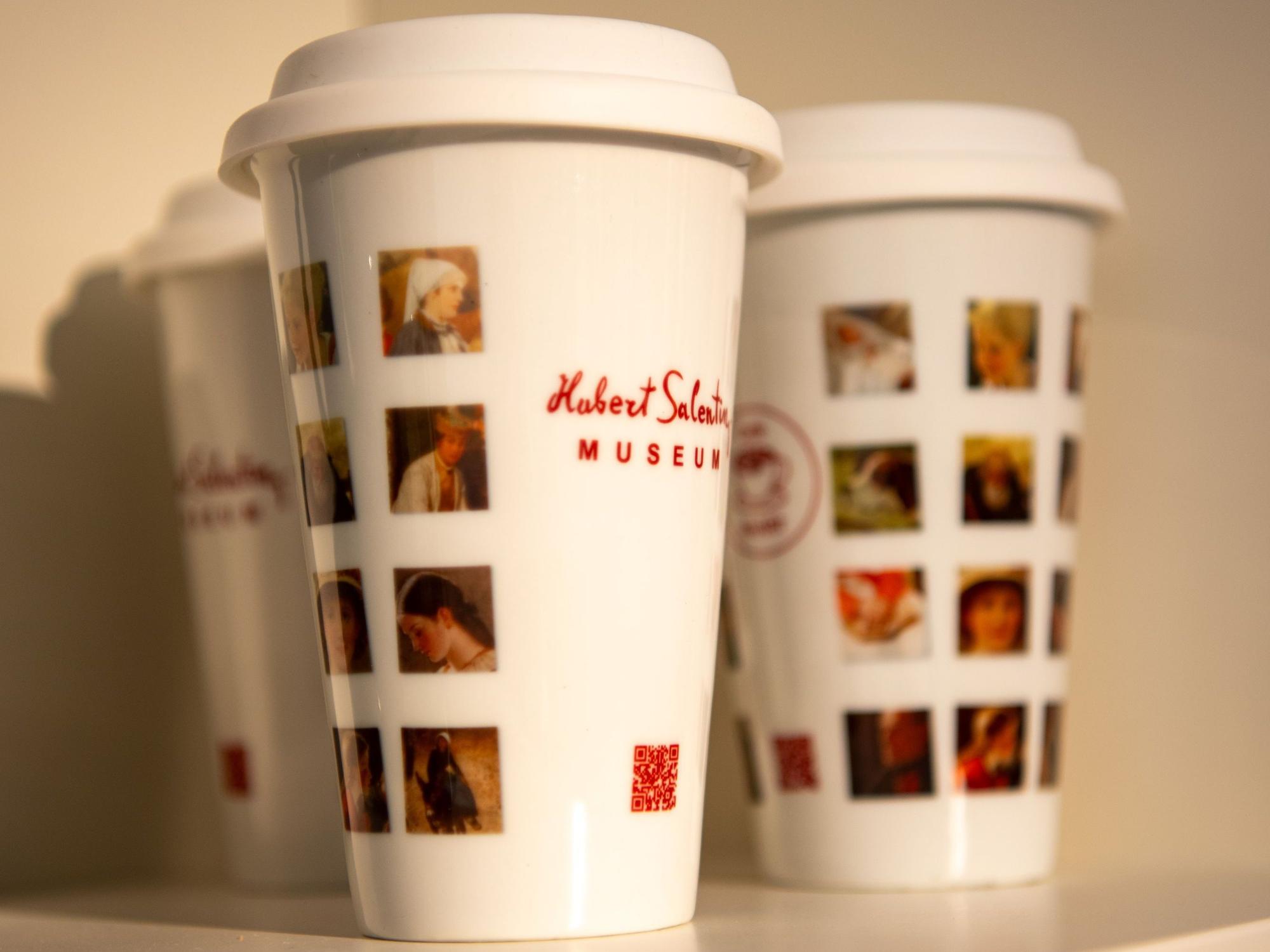 Mehrweg-Kaffeebecher mit dem Aufdruck „Hubert Salentin Museum“ und verschiedenen Bildausschnitten von Salentins Gemälden stehen in einem Regal.