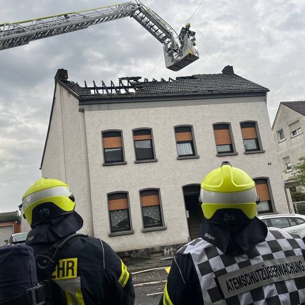 Feuerwehrleute stehen vor einem Mehrfamilienhaus, über dem ausgebrannten Dach schwebt der Korb der Drehleiter.