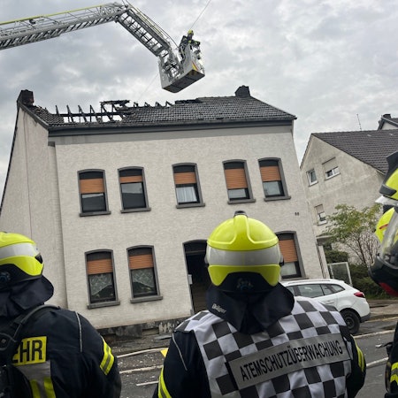 Feuerwehrleute stehen vor einem Mehrfamilienhaus, über dem ausgebrannten Dach schwebt der Korb der Drehleiter.