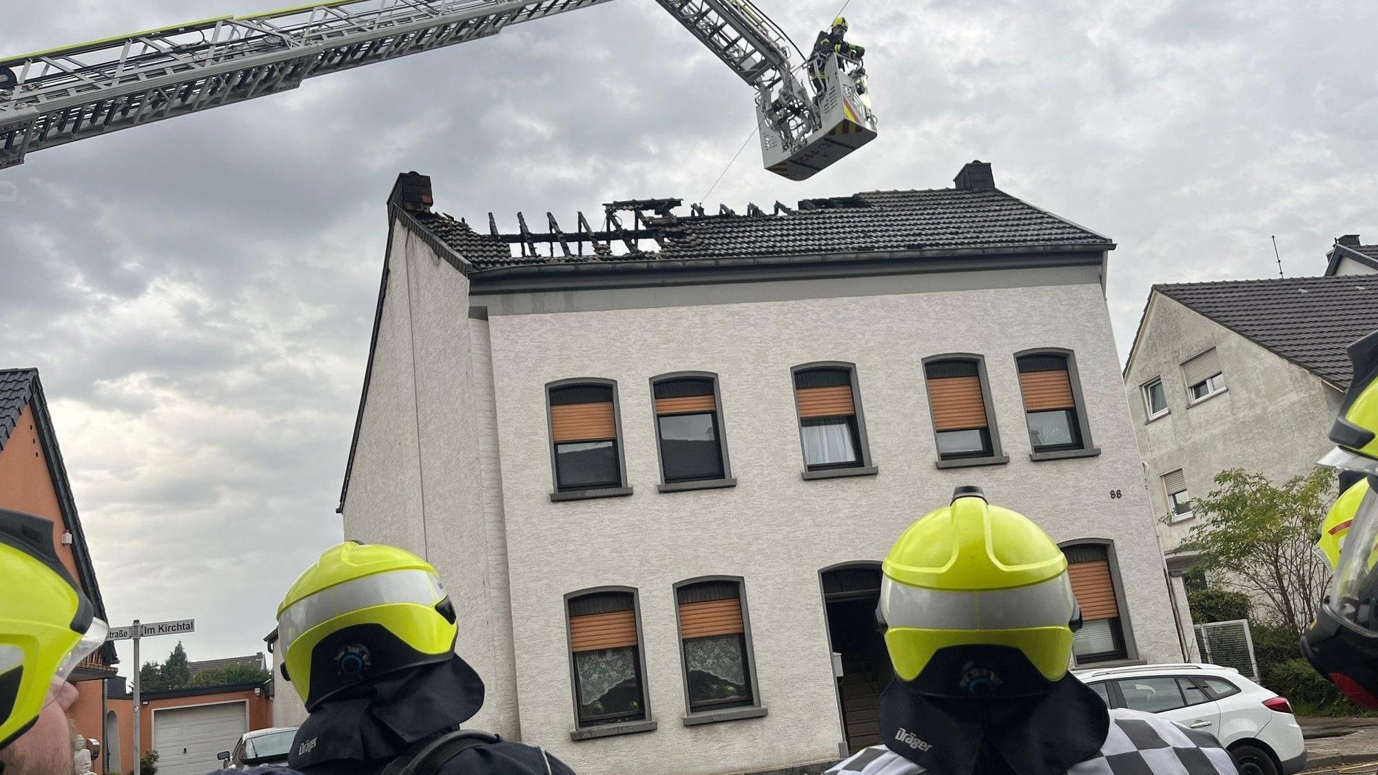 Feuerwehrleute stehen vor einem Mehrfamilienhaus, über dem ausgebrannten Dach schwebt der Korb der Drehleiter.