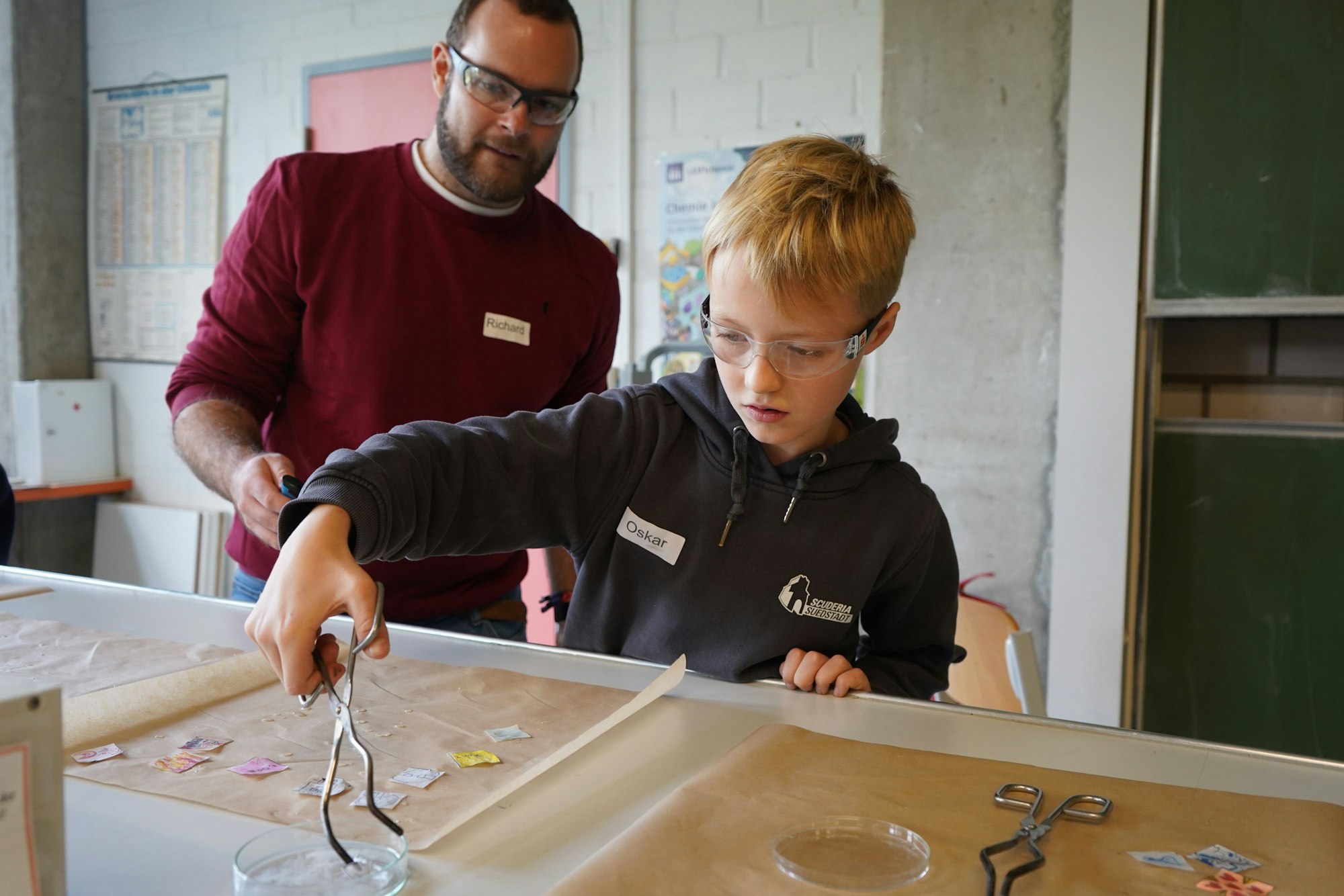 Das Bild zeigt ein Kind, das mit einer Zange ein Stück Papier in eine Flüssigkeit taucht.