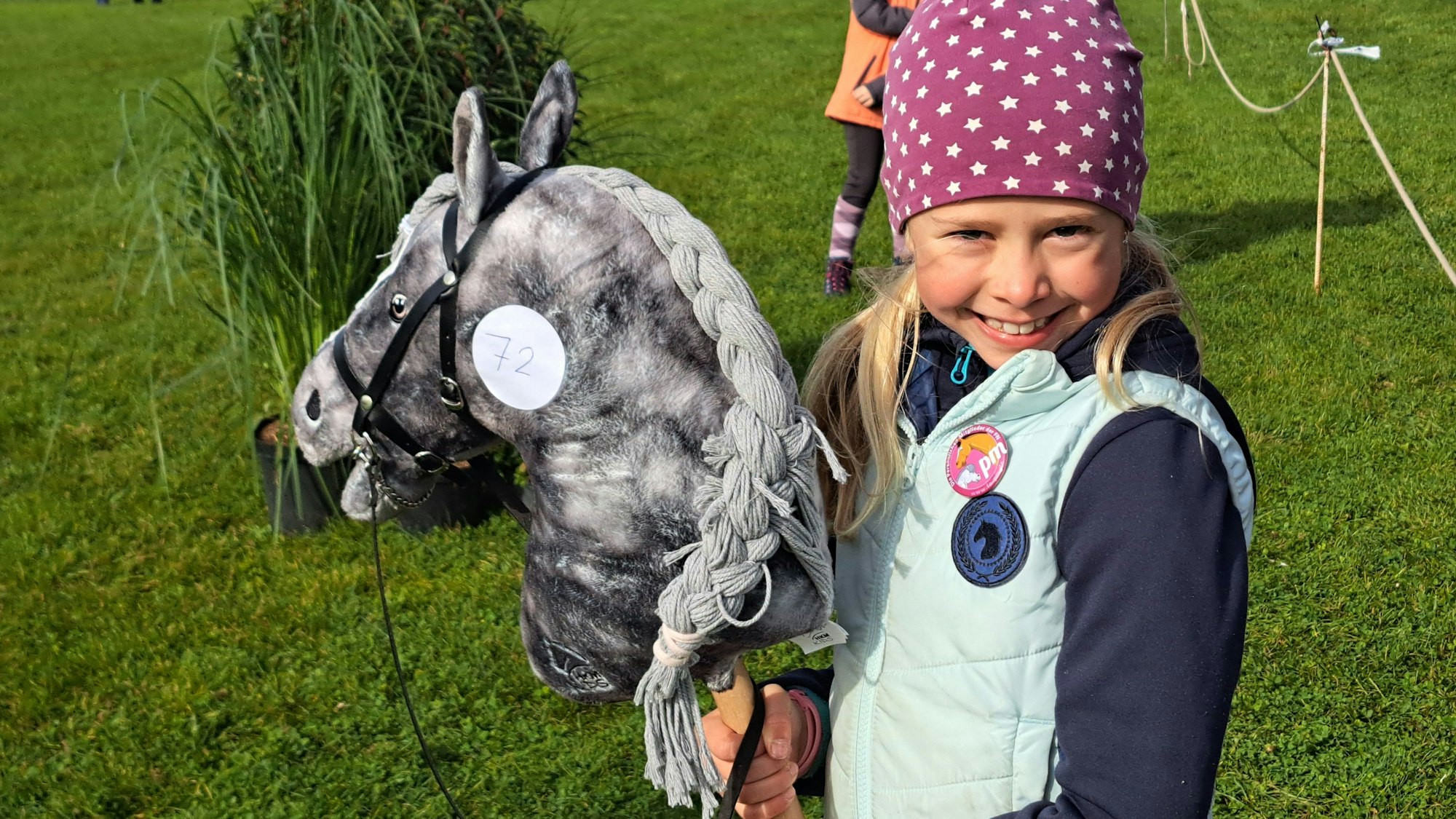 Das Bild zeigt das Mädchen, das in die Kamera lächelt. In der Hand hält es ein Steckenpferd.