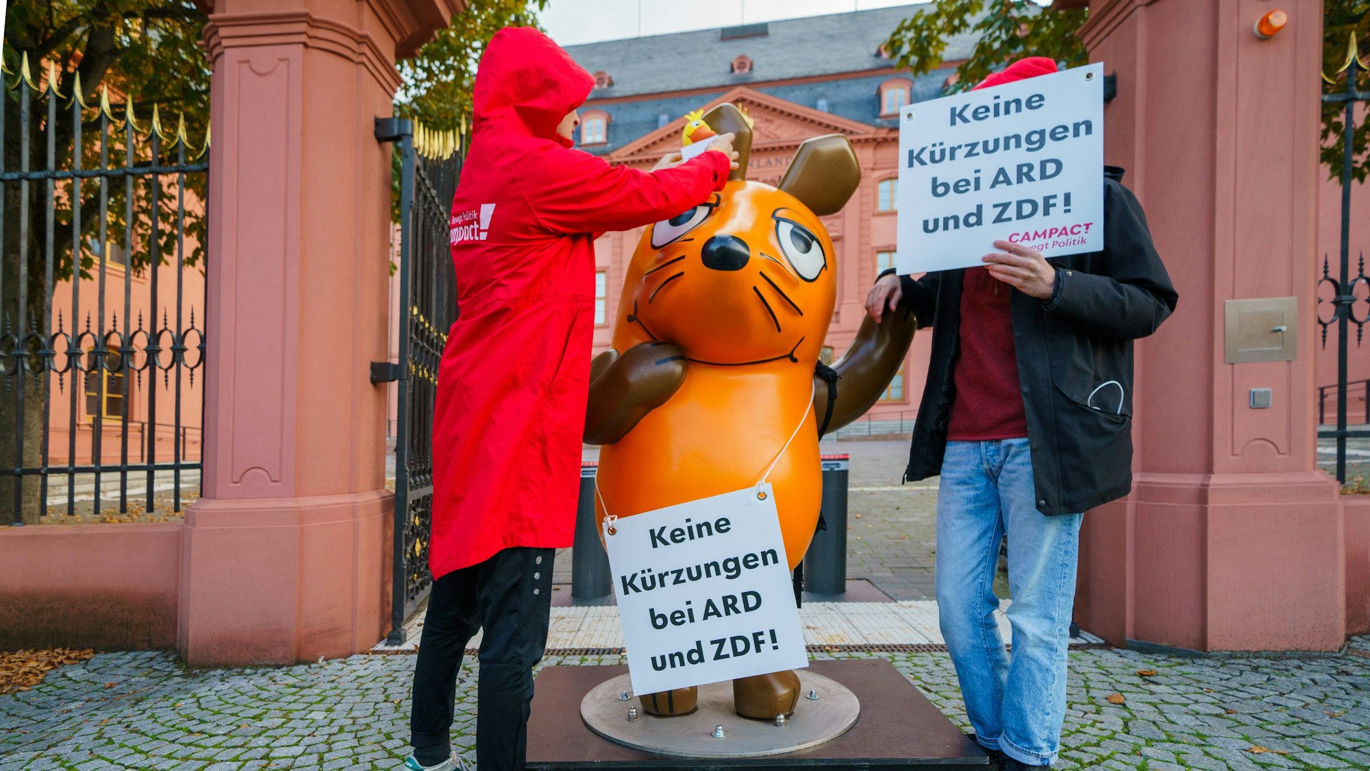 Die Figur der Maus aus der „Sendung mit der Maus“ steht zwischen Aktivisten von Campact vor dem Landtag Rheinland-Pfalz, die Schilder mit der Aufschrift «Keine Kürzungen bei ARD und ZDF!» halten