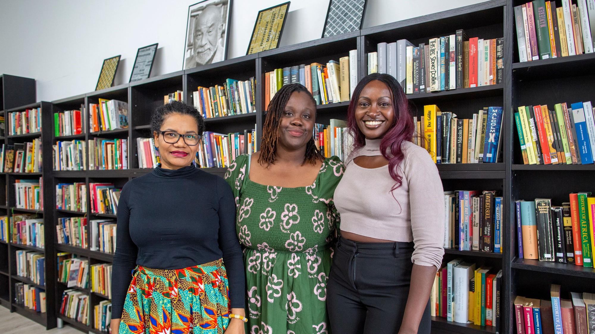 Glenda Obermuller (v.l.n.r.), N'joula Baryoh und Sängerin Naiyango stehen vor gefüllten Bücherregalen.