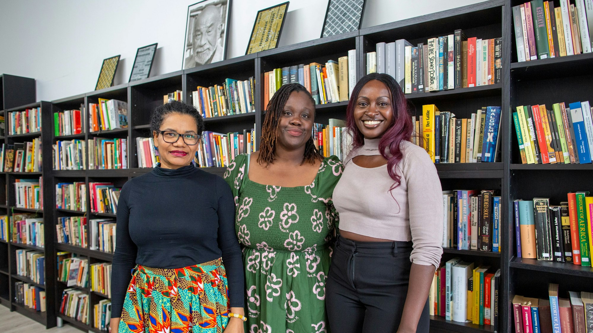 Glenda Obermuller (v.l.n.r.), N'joula Baryoh und Sängerin Naiyango stehen vor gefüllten Bücherregalen.