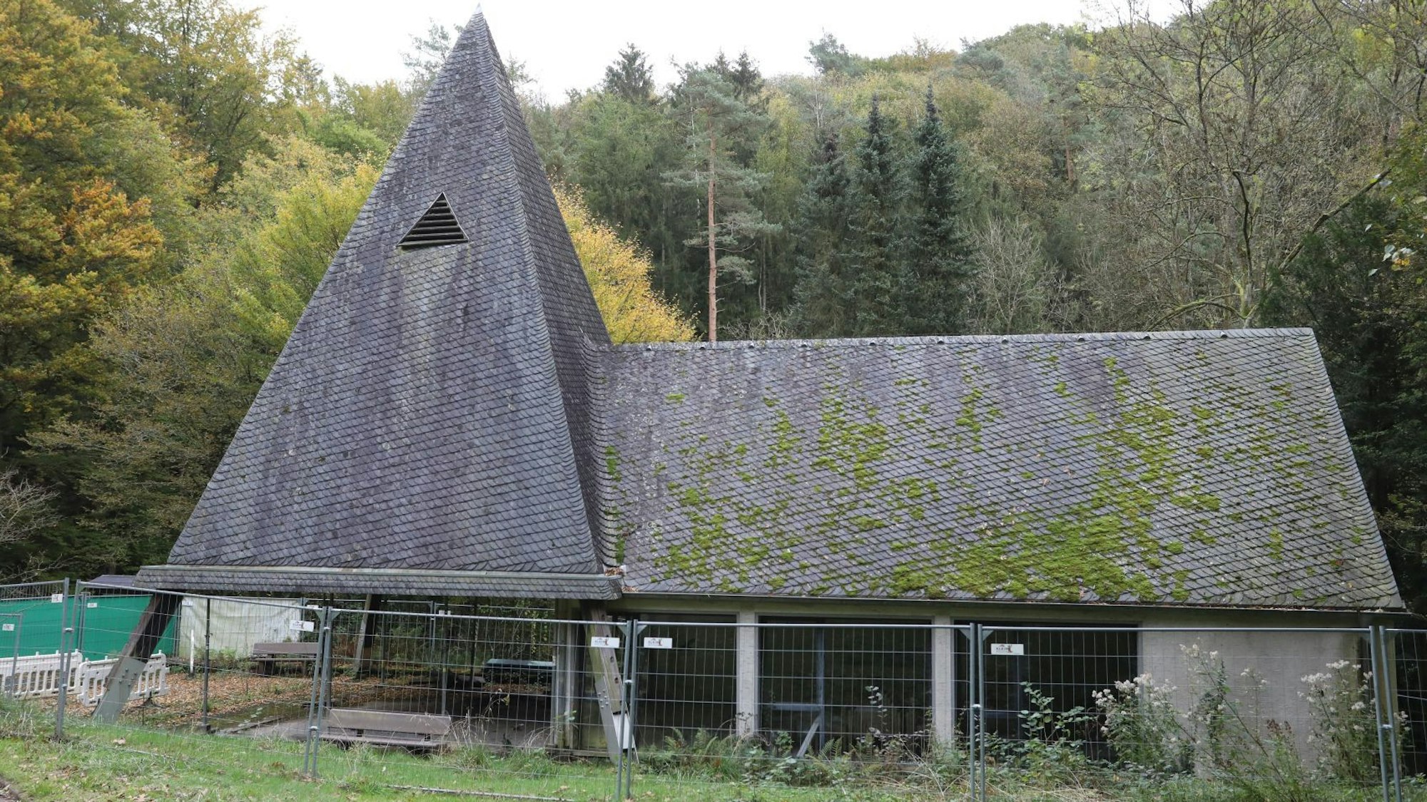 Ein Bauzaun umgibt eine Kapelle, auf deren Dach zum Teil grünes Moos wächst.
