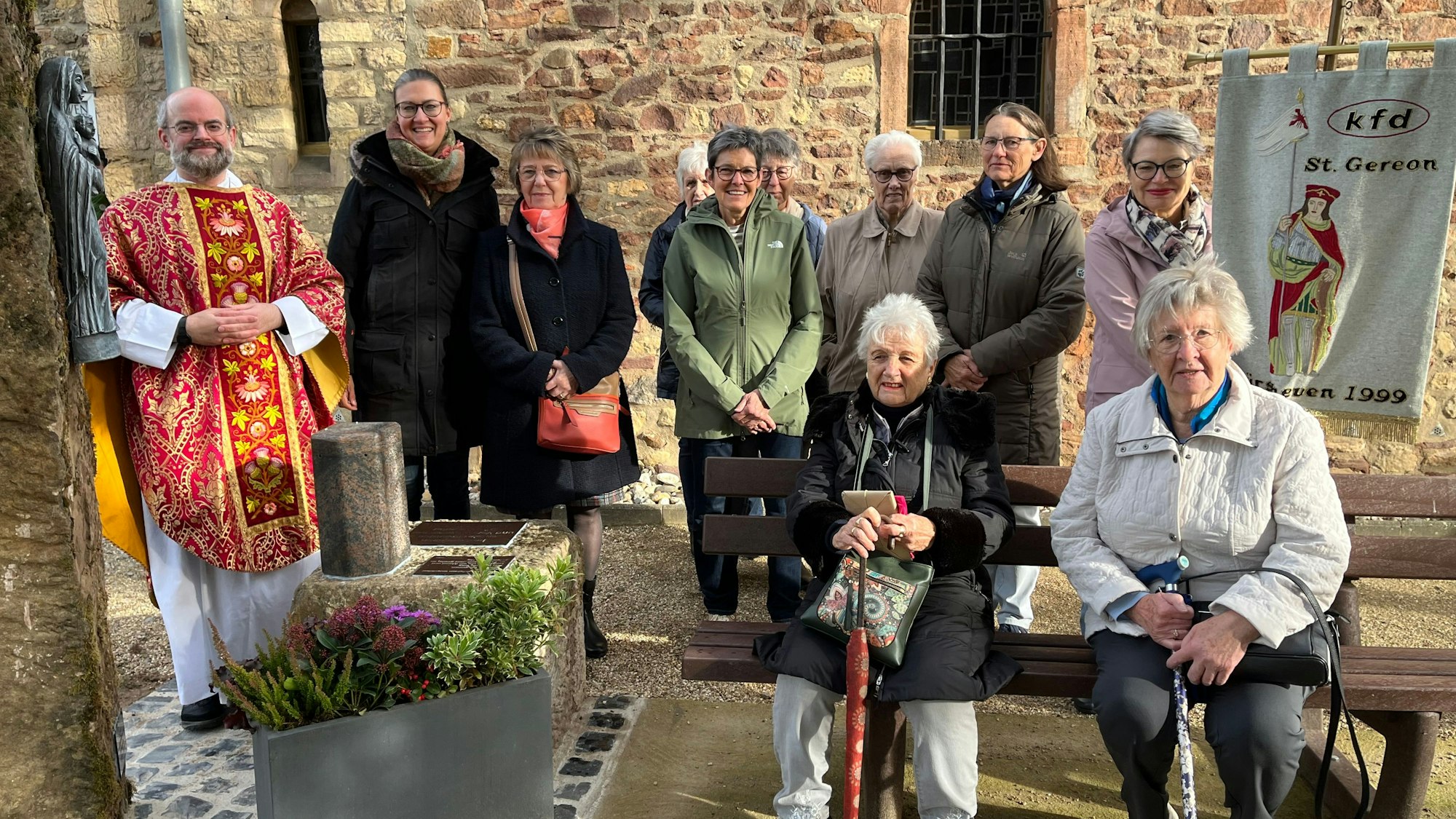 Eine Gruppe Frauen und ein Pastor sitzen und stehen vor einer Kirchenmauer. Links im Bild ist eine Marienfigur an einer Stele zu sehen.