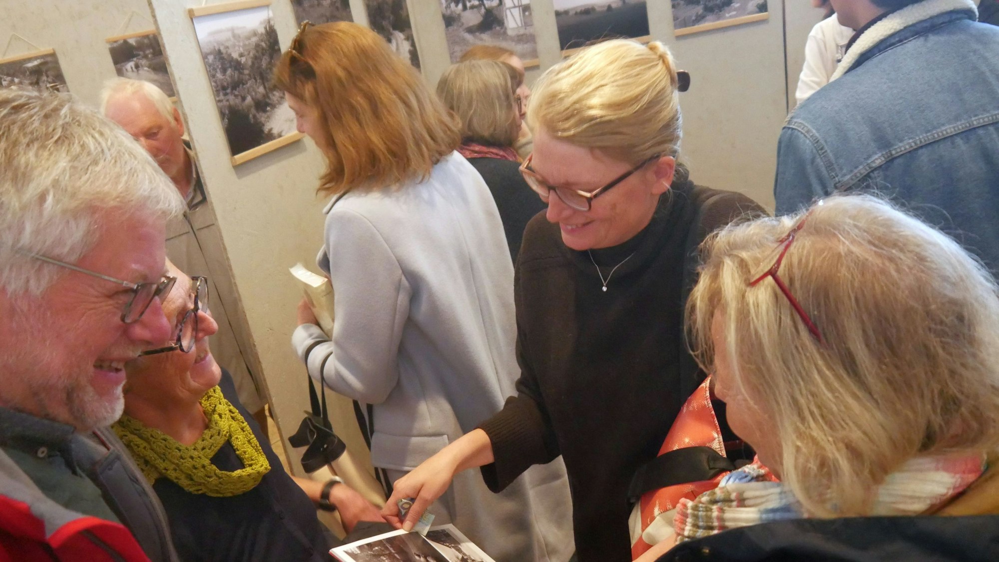 Menschen stehen in einer Ausstellung mit Schwarz-Weiß-Fotografien zusammen, eine blonde Frau mit Brille blättert in einem Katalog.