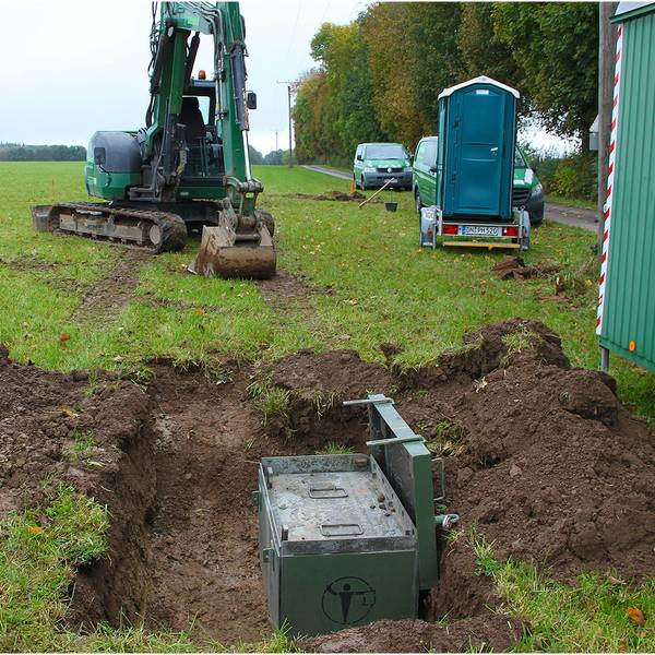 Auf einer Wiese mit einem ausgehobenen Loch, in der eine grüne Kiste steht, stehen ein Minibagger, ein mobiles WC und der Bauwagen eines Kampfmittelräumdienstes.