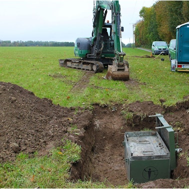 Auf einer Wiese mit einem ausgehobenen Loch, in der eine grüne Kiste steht, stehen ein Minibagger, ein mobiles WC und der Bauwagen eines Kampfmittelräumdienstes.