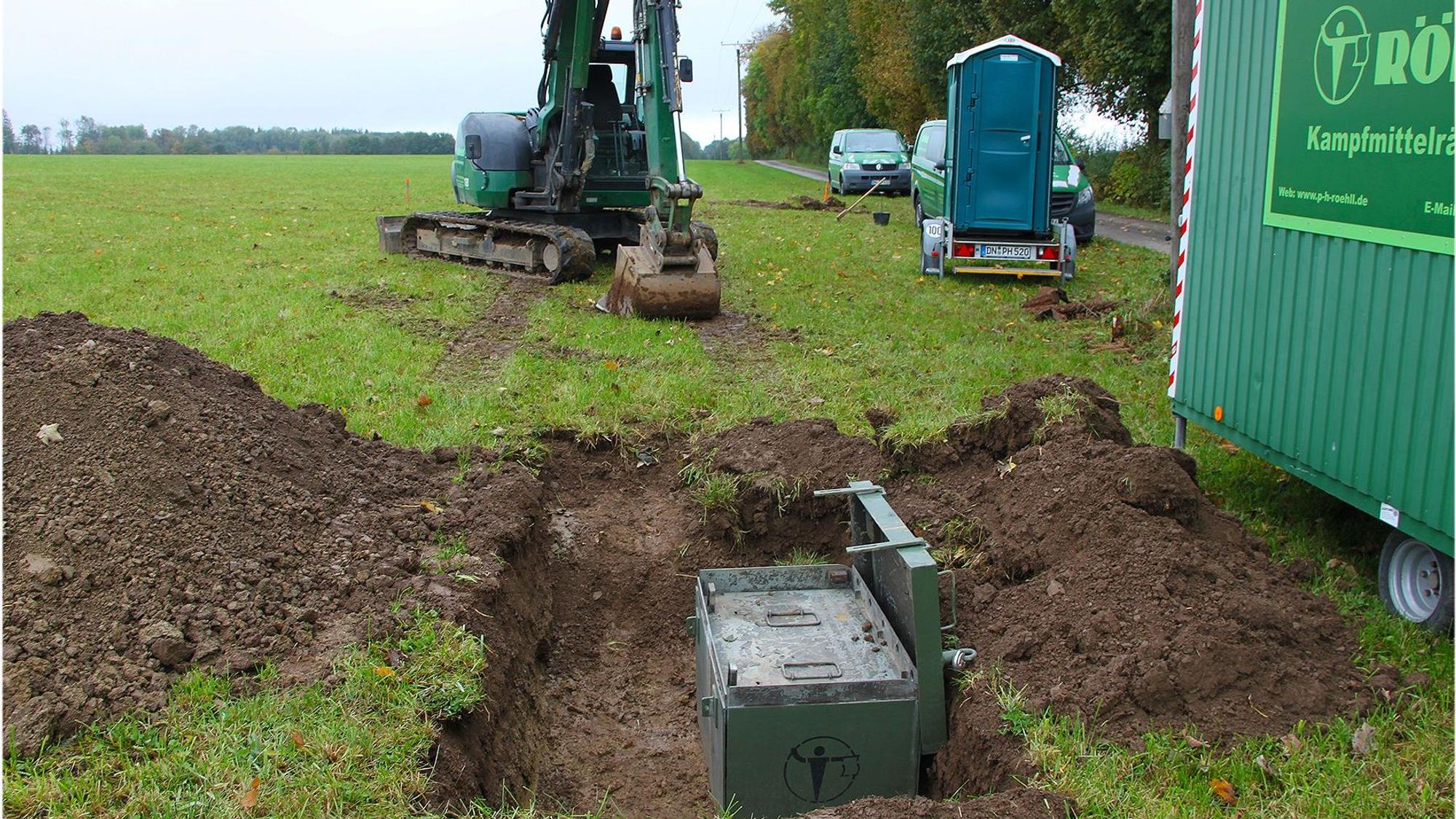 Auf einer Wiese mit einem ausgehobenen Loch, in der eine grüne Kiste steht, stehen ein Minibagger, ein mobiles WC und der Bauwagen eines Kampfmittelräumdienstes.