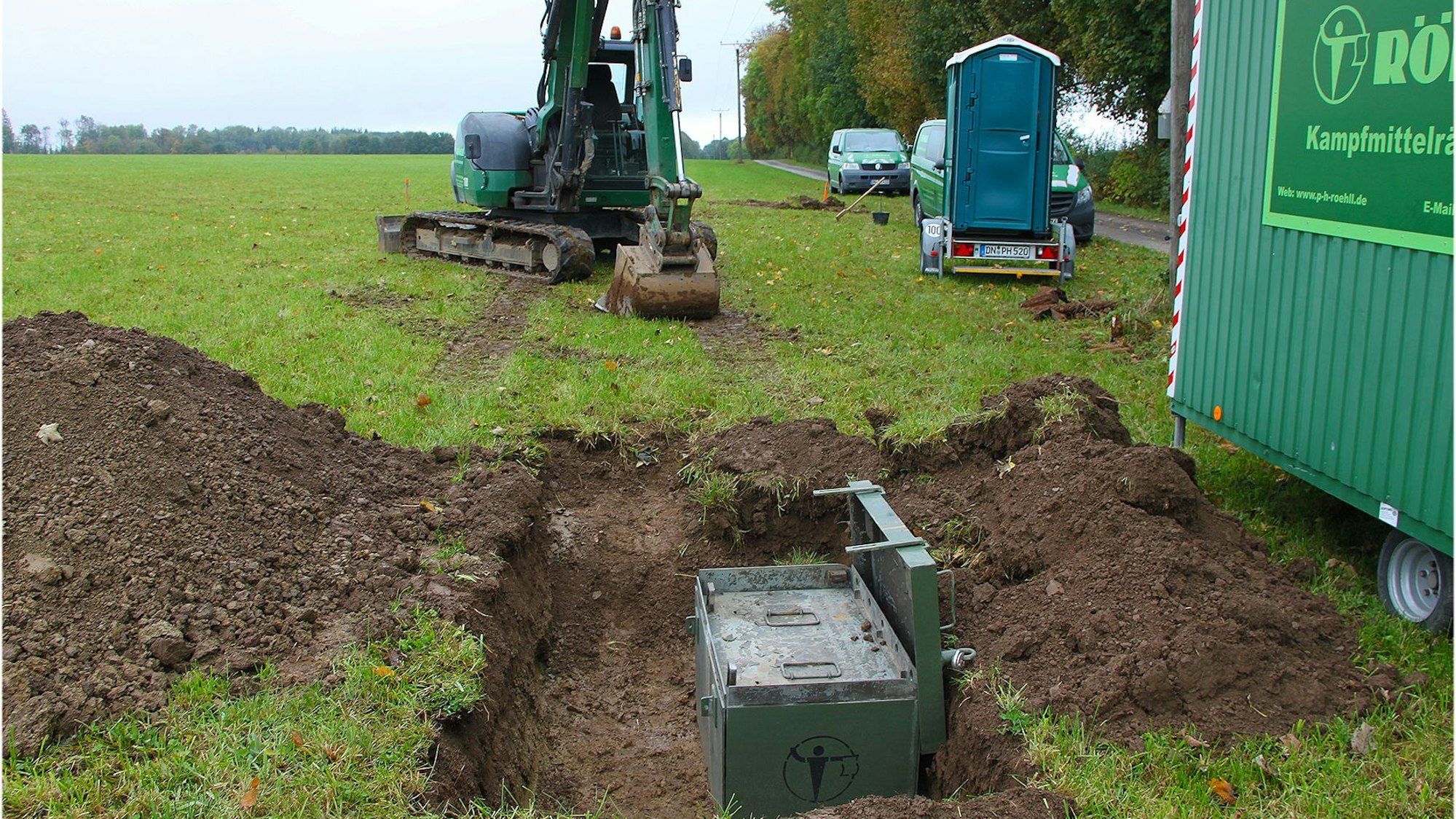 Auf einer Wiese mit einem ausgehobenen Loch, in der eine grüne Kiste steht, stehen ein Minibagger, ein mobiles WC und der Bauwagen eines Kampfmittelräumdienstes.