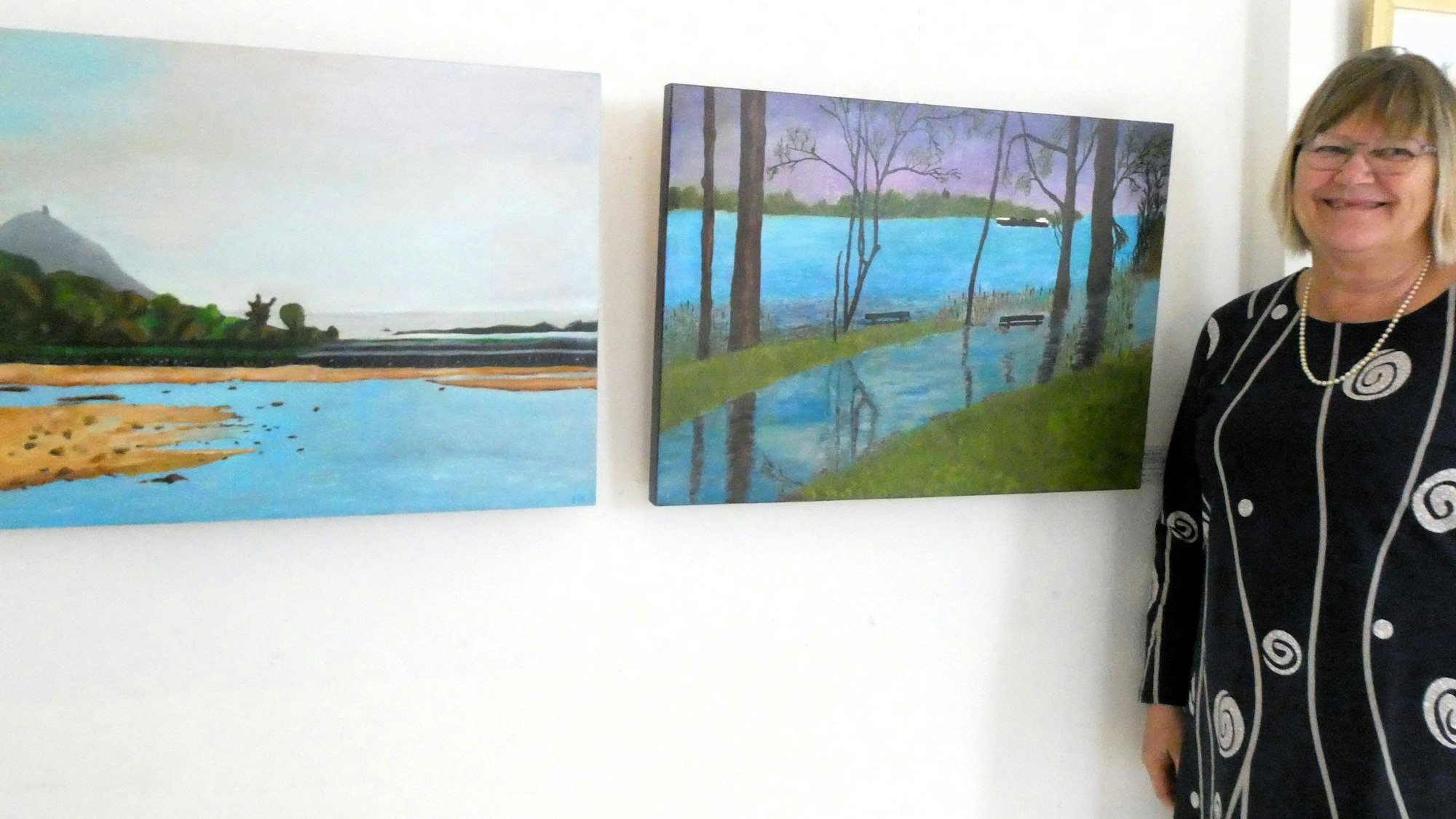 Eine Frau neben zwei Kunstwerken, von denen eines den Rhein und den Drachenfels zeigt.