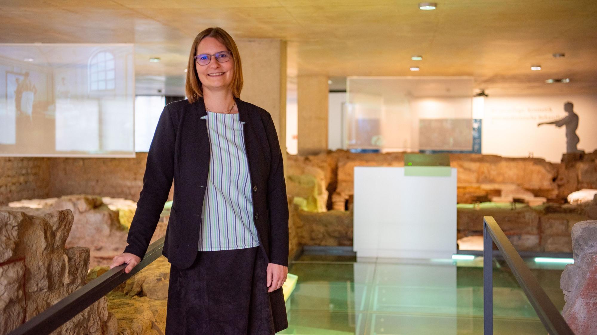 Dr. Silva Bruder lehnt in gestreifter Bluse und dunkelblauem Blazer an einem Geländer in der Ausstellung des Museums für Badekultur. Um sie herum sind die archäologischen Überreste einer antiken Thermen-Anlage zu sehen.