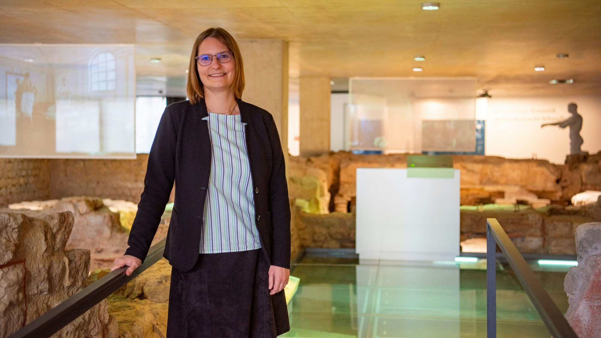 Dr. Silva Bruder lehnt in gestreifter Bluse und dunkelblauem Blazer an einem Geländer in der Ausstellung des Museums für Badekultur. Um sie herum sind die archäologischen Überreste einer antiken Thermen-Anlage zu sehen.