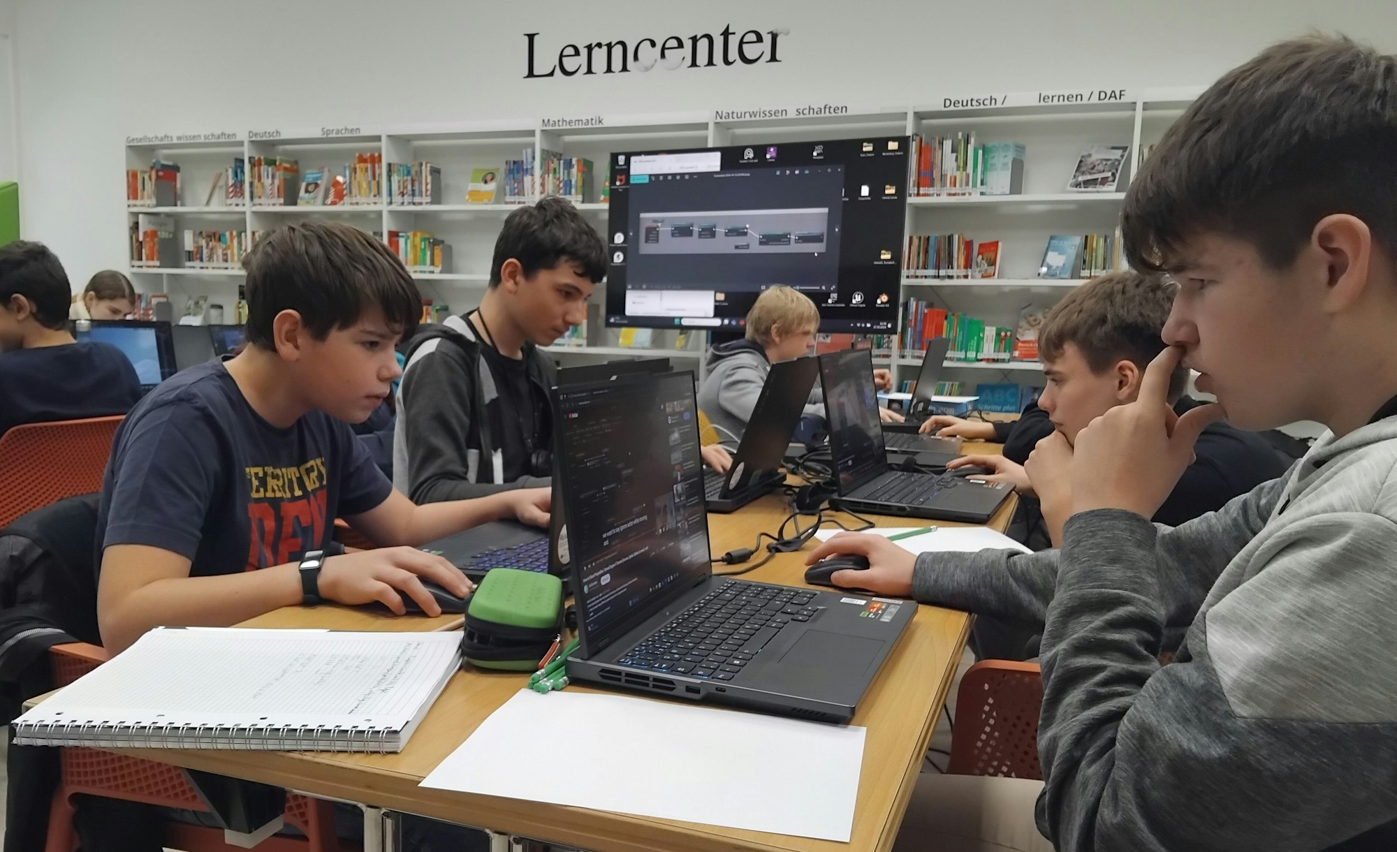 Fünf Jungen im Alter von 13 bis 15 Jahren sitzen vor aufgeklappten Laptops an einem Tisch und schauen konzentriert. Neben ihnen liegen Notizblöcke. Im Hintergrund sieht man weitere Jugendliche und einen großen Bildschirm, außerdem die Bücherregale der Bibliothek. "Lerncenter" steht in schwarzen Lettern an der Wand.