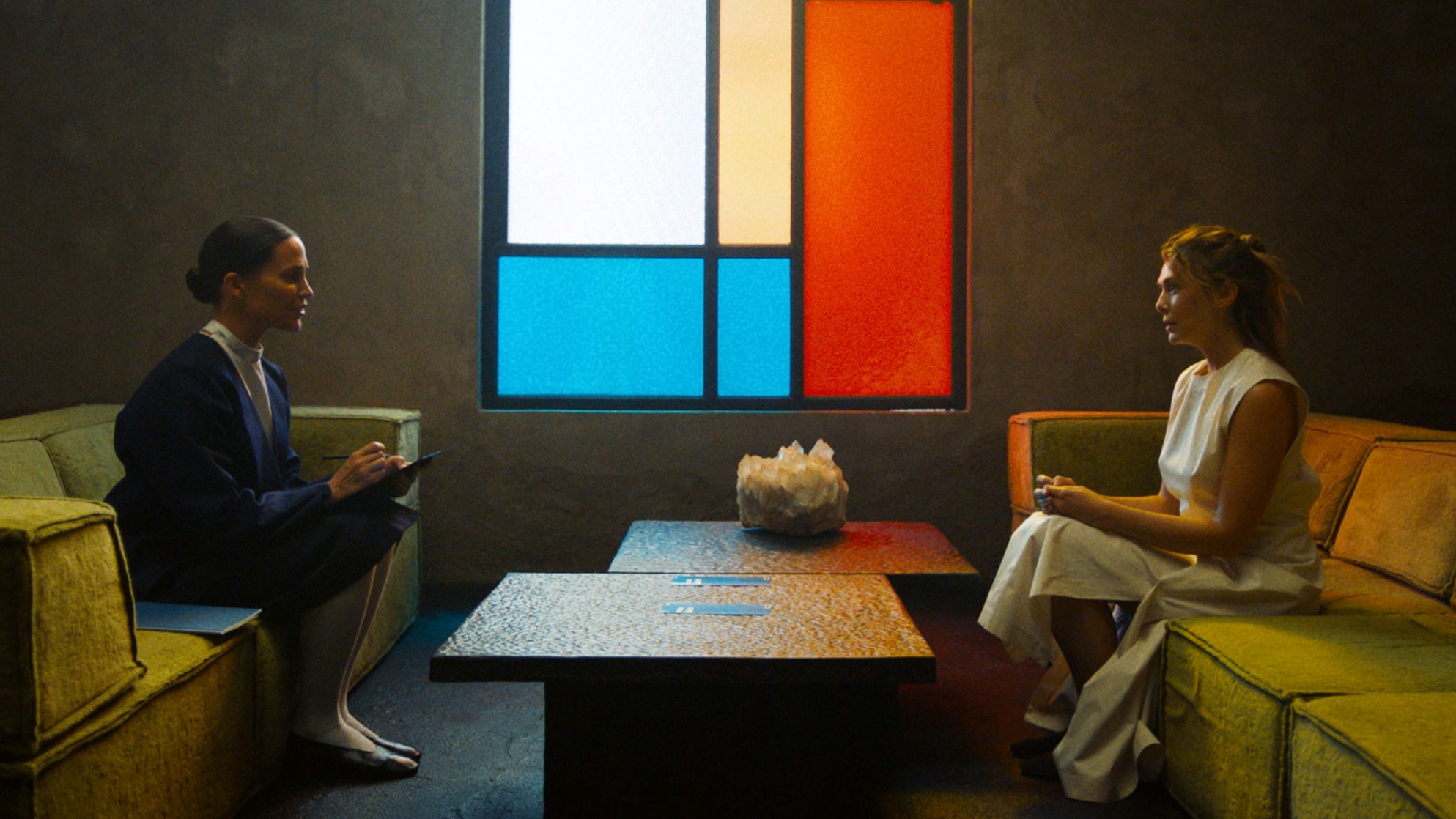 Zwei Frauen sitzen sich mit sehr viel Abstand gegenüber, wobei eine von ihnen ein Klemmbrett in den Händen hält und sich Notizen macht.