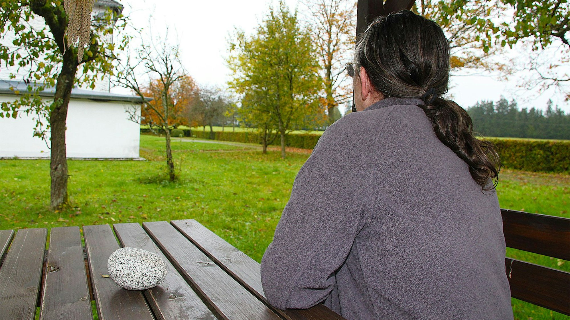 Eine Frau sitzt auf einer Bank und schaut in die Landschaft mit Wiese und Bäumen.