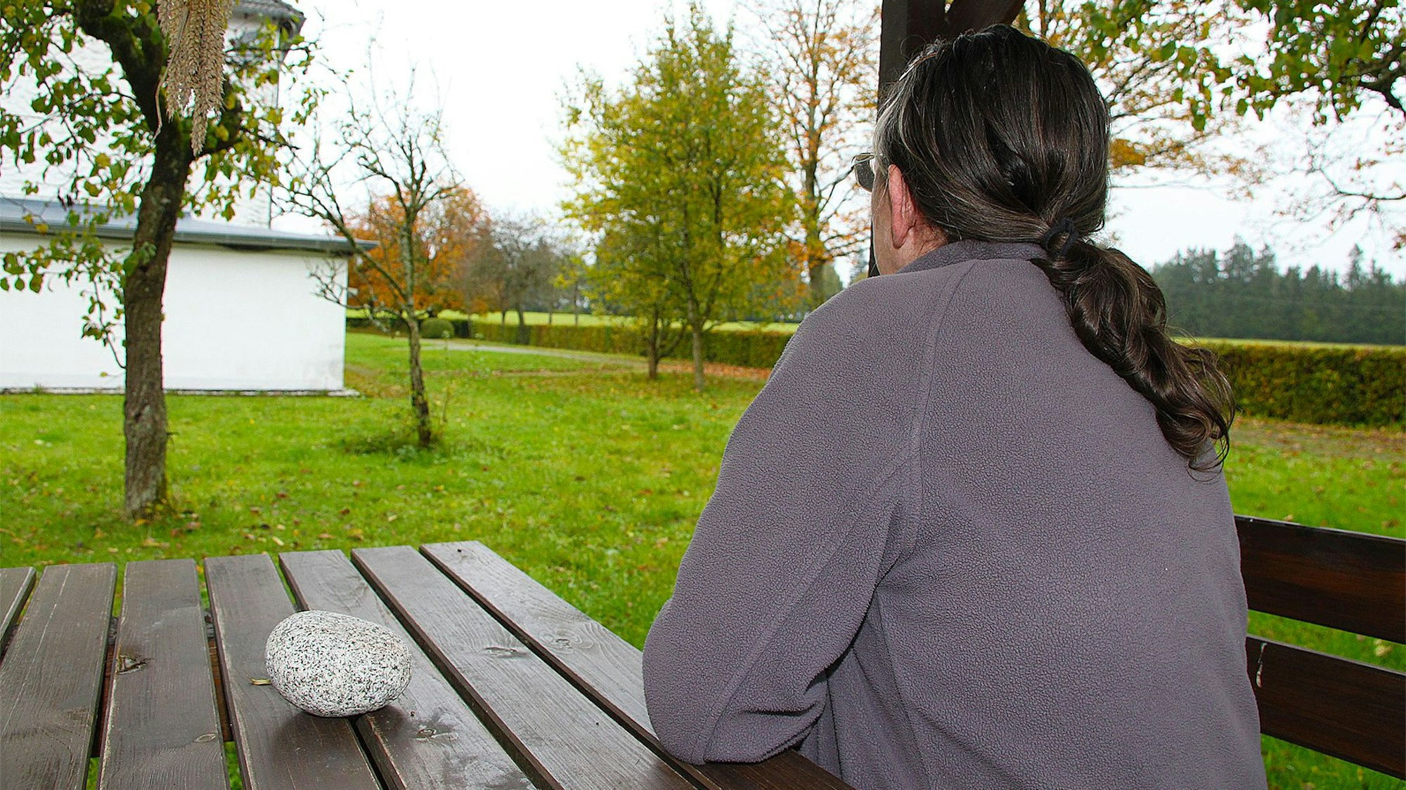 Eine Frau sitzt auf einer Bank und schaut in die Landschaft mit Wiese und Bäumen.
