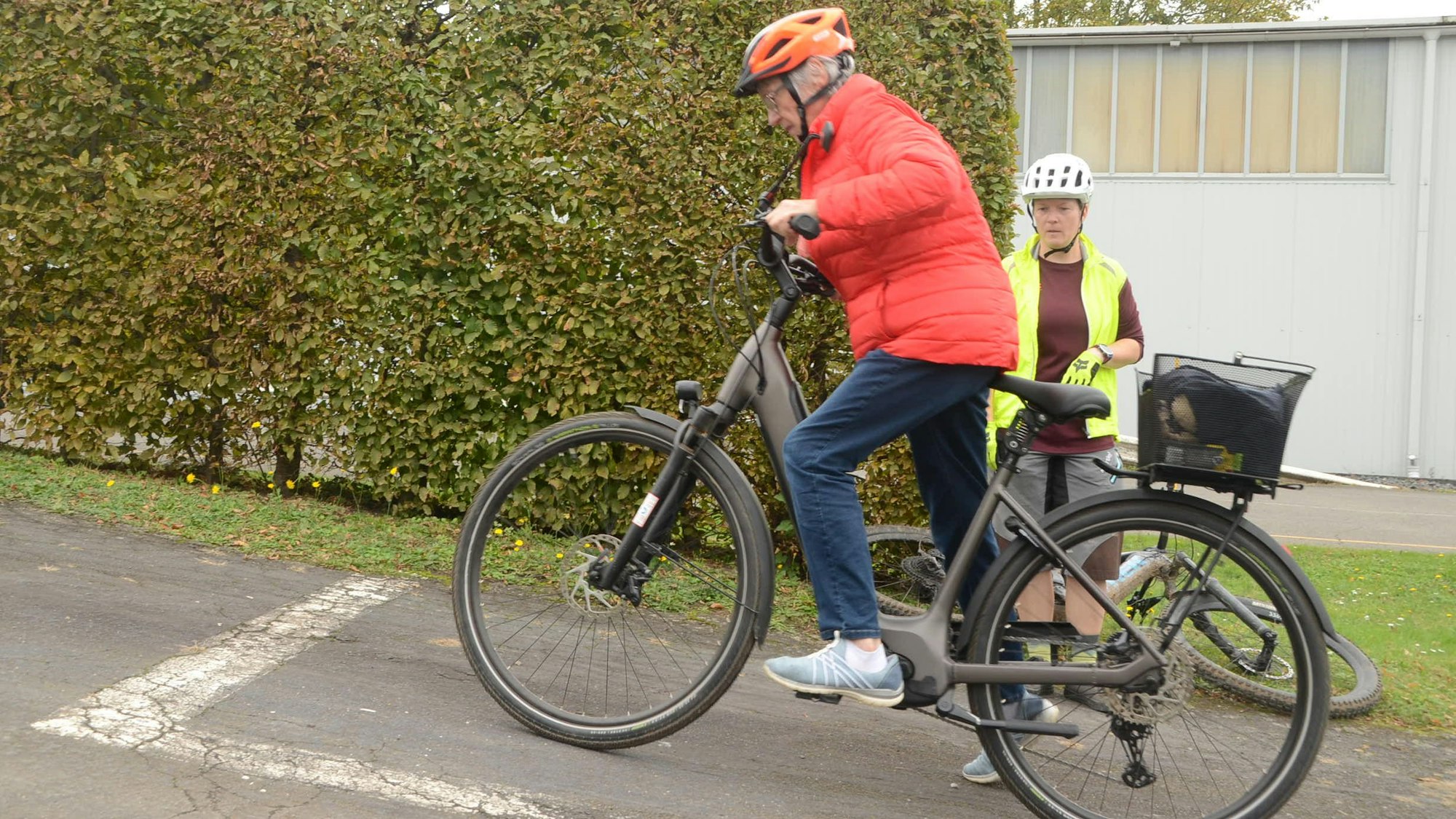 Eine Frau in einer roten Windjacke und mit Fahrradhelm schiebt ihr Pedelec an einer Steigung an.