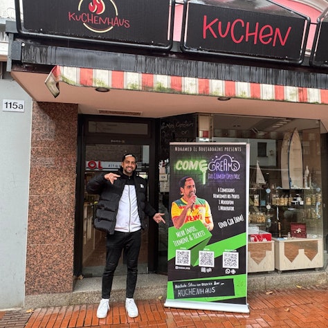 Auf dem Bild ist der Comedian vor einem Veranstaltungsplakat zu sehen.