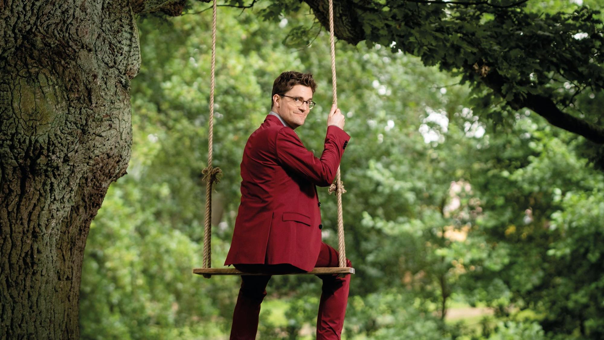 Das Bild zeigt Bodo Wartke auf einer Schaukel, sitzend, Blick über die Schulter. Er trägt einen roten Anzug.