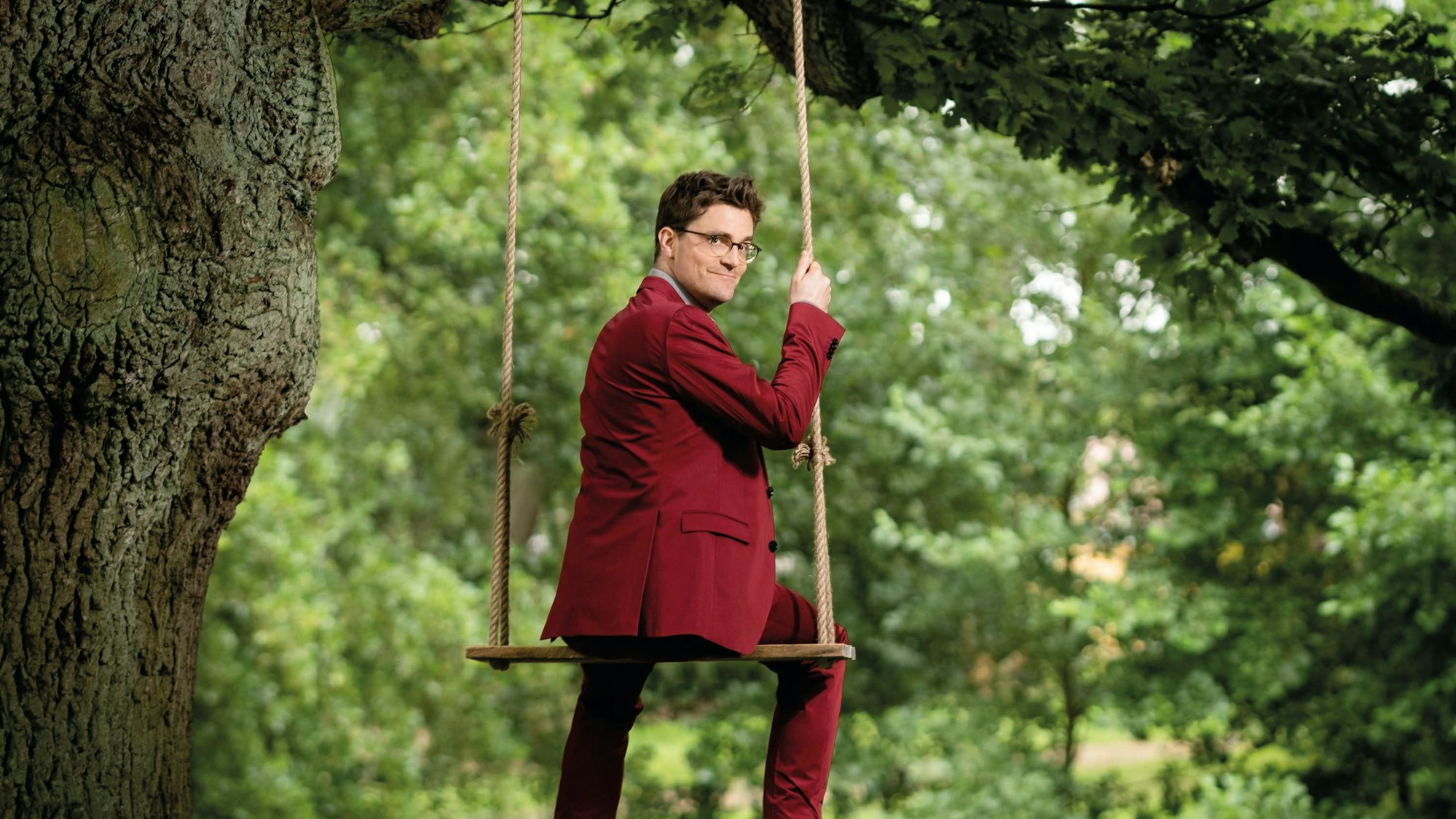 Das Bild zeigt Bodo Wartke auf einer Schaukel, sitzend, Blick über die Schulter. Er trägt einen roten Anzug.