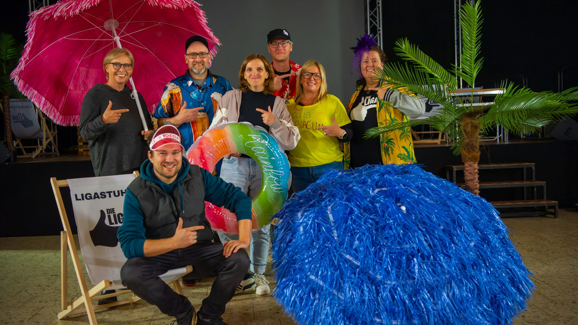 17.10.2024 Zülpich-Niederelvenich. Vier Frauen und drei Männer stehen in bunten Hawaihemden und mit bunten Sonnenschirmen vor einer Bühne. Isa Oestmann (v.l.), Jens Bongard, Stephanie Schneider, Andreas Krantz, Birte Karsten, Eva Lebertz und Julius Esser (sitzend) proben für die Sitzungen der Liga der alternativen Karnevalisten.
