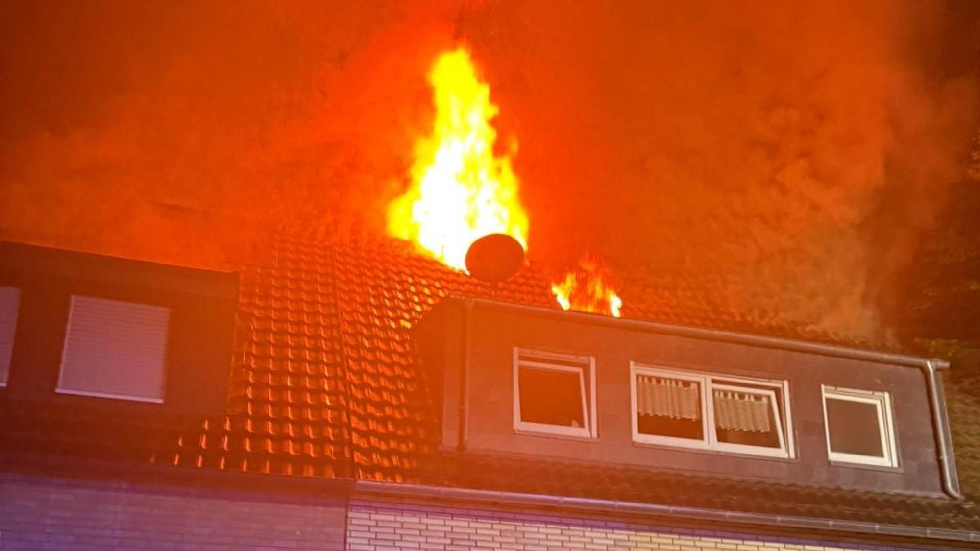Das Foto zeigt den Dachstuhl mit Gaube eines Mehrfamilienhauses. Aus dem Dach schlagen hohe Flammen.