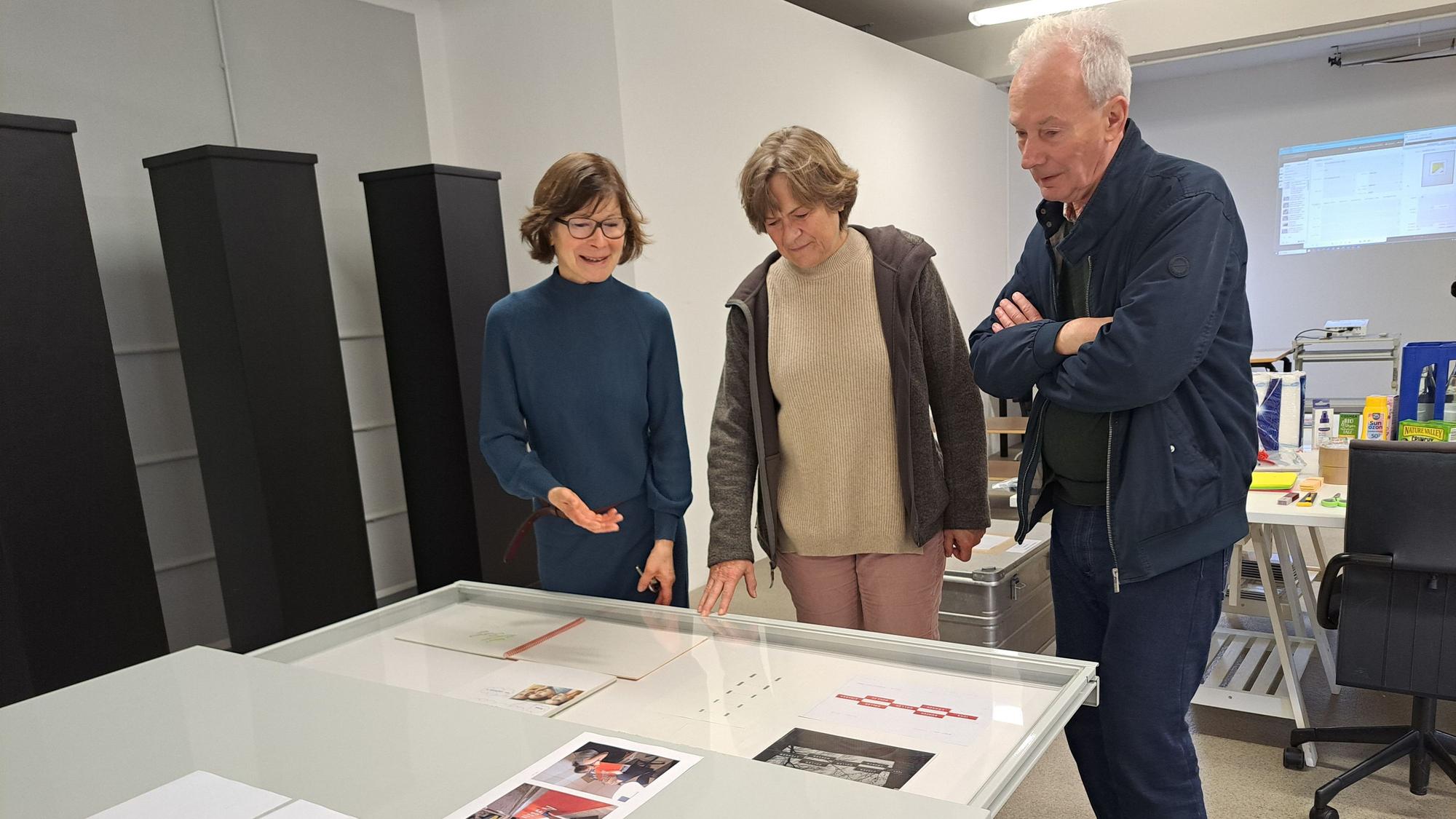 Zwei Frauen und ein Mann schauen sich Kunstwerke, die vor ihnen liegen, an.