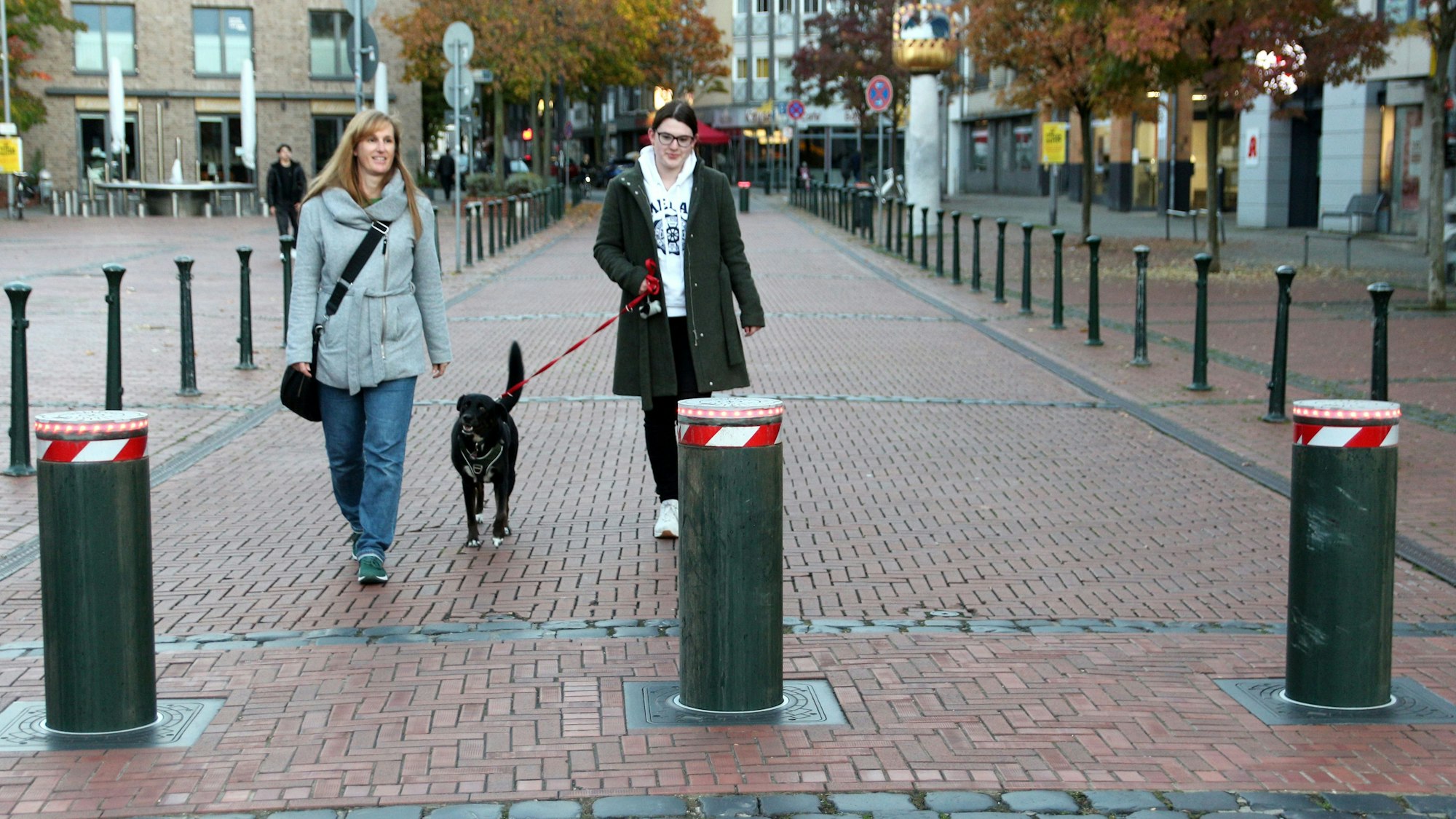Zwei Frauen gehen mit einem Hund spazieren, vor ihnen sperren Poller den Straßenabschnitt ab.