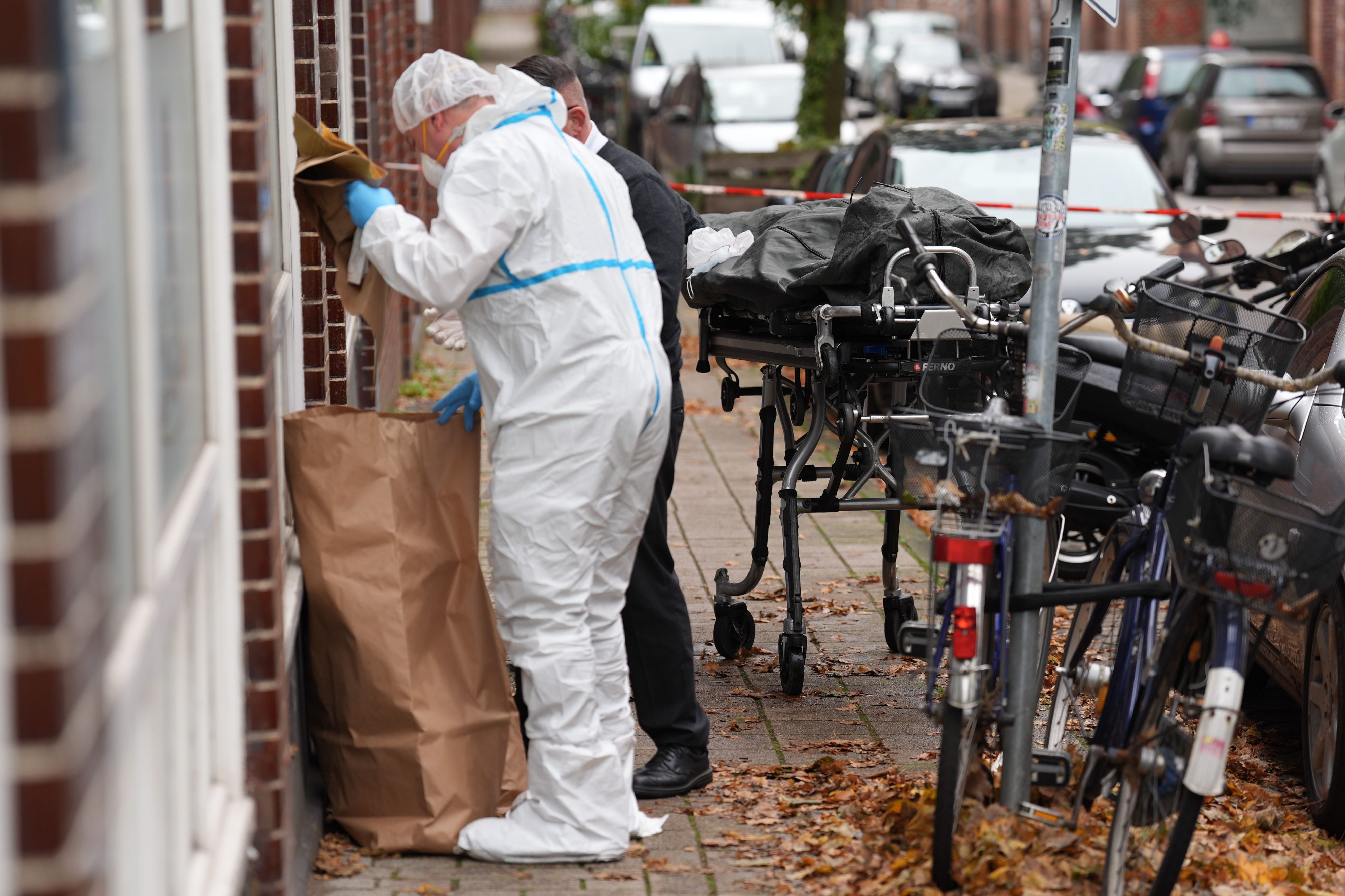 Ein Toter liegt in einem Leichensack (r) auf einer Trage neben einem Mitarbeiter der Kriminaltechnik und einem Bestatter vor einem Mehrfamilienhaus.