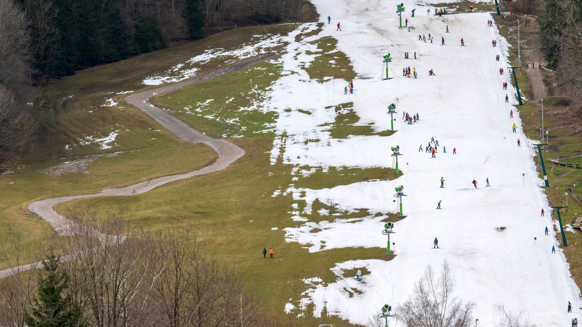04.01.2024, Bayern, Brauneck: Skifahrer nutzen am Vormittag die beschneite Fläche im Talbereich für ihr kühles Wintervergnügen. Links zieht sich ein Feldweg durch eine grüne Wiese.