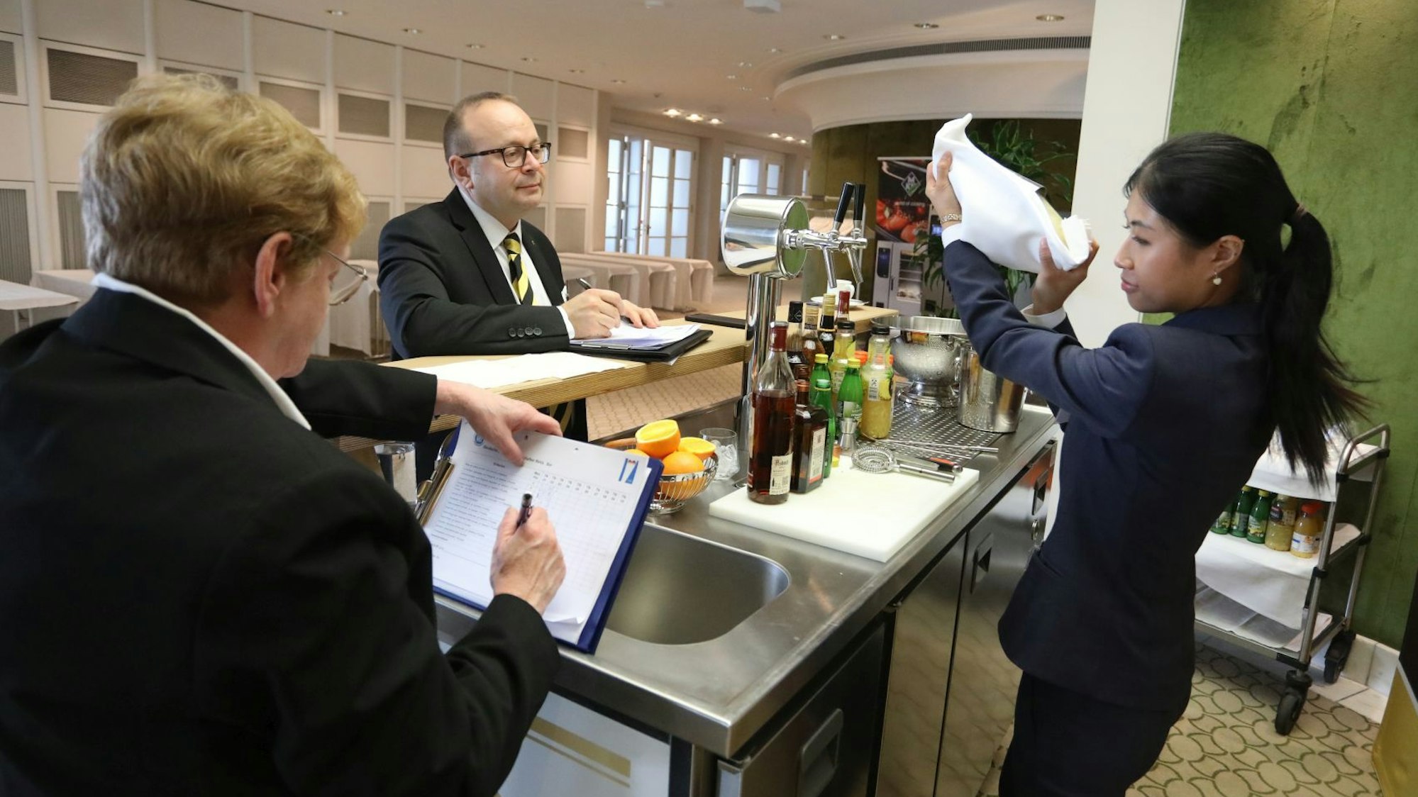 Eine junge Frau mixt an einer kleinen Bar ein Getränk, zwei Jurorfn machen sich Notizen.