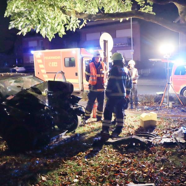Zu sehen ist ein Unfall bei Nacht, ein Auto ist beschädigt, Feuerwehrleute stehen daneben, ein Krankenwagen steht im Hintergrund.