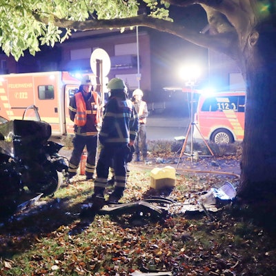 Zu sehen ist ein Unfall bei Nacht, ein Auto ist beschädigt, Feuerwehrleute stehen daneben, ein Krankenwagen steht im Hintergrund.