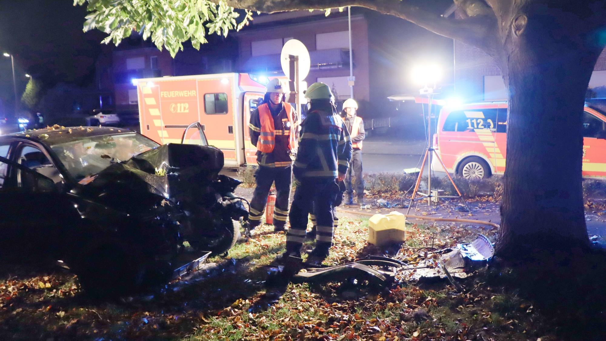 Zu sehen ist ein Unfall bei Nacht, ein Auto ist beschädigt, Feuerwehrleute stehen daneben, ein Krankenwagen steht im Hintergrund.