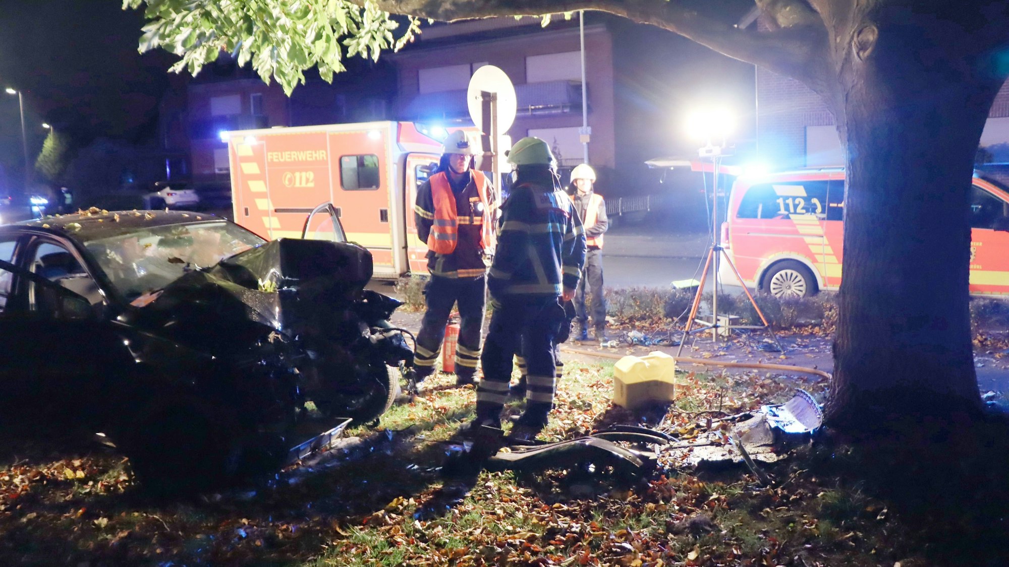 Zu sehen ist ein Unfall bei Nacht, ein Auto ist beschädigt, Feuerwehrleute stehen daneben, ein Krankenwagen steht im Hintergrund.