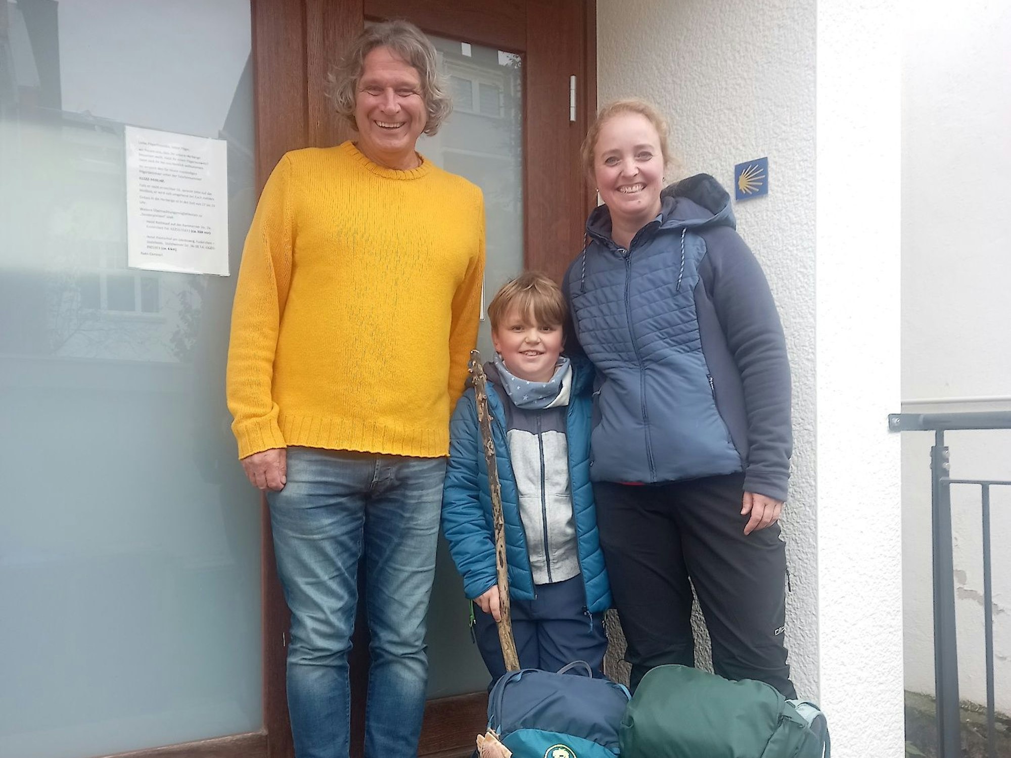 Zwei Erwachsene und ein Kind stehen vor einem Hauseingang und grinsen in die Kamera.